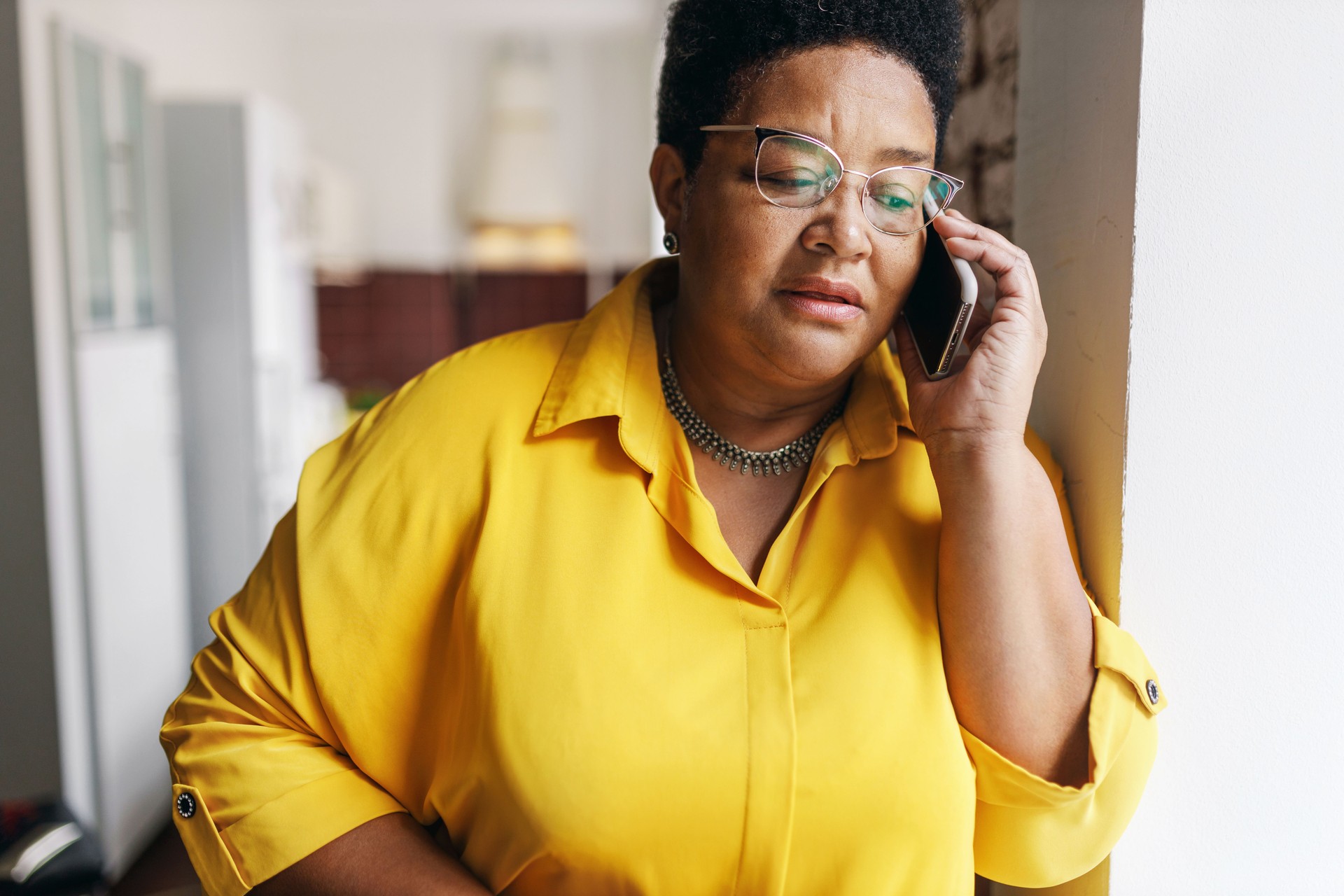 Serious african american senior woman in glasses and yellow shirt talking on phone with serious worried facial expression, receiving bad news from her doctor or relatives, leaning on wall